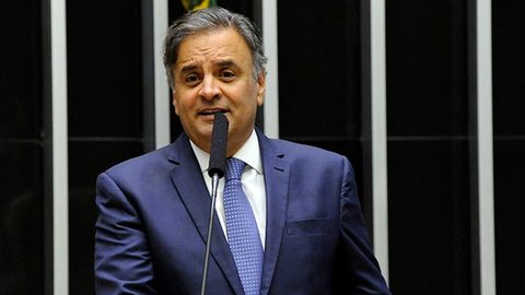 Fotografia. Aécio Neves, de terno azul e gravata, fala ao microfone no plenário da Câmara dos Deputados.