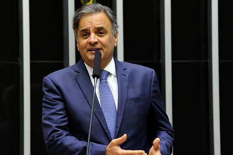 Fotografia. Aécio Neves, de terno azul e gravata, fala ao microfone no plenário da Câmara dos Deputados.