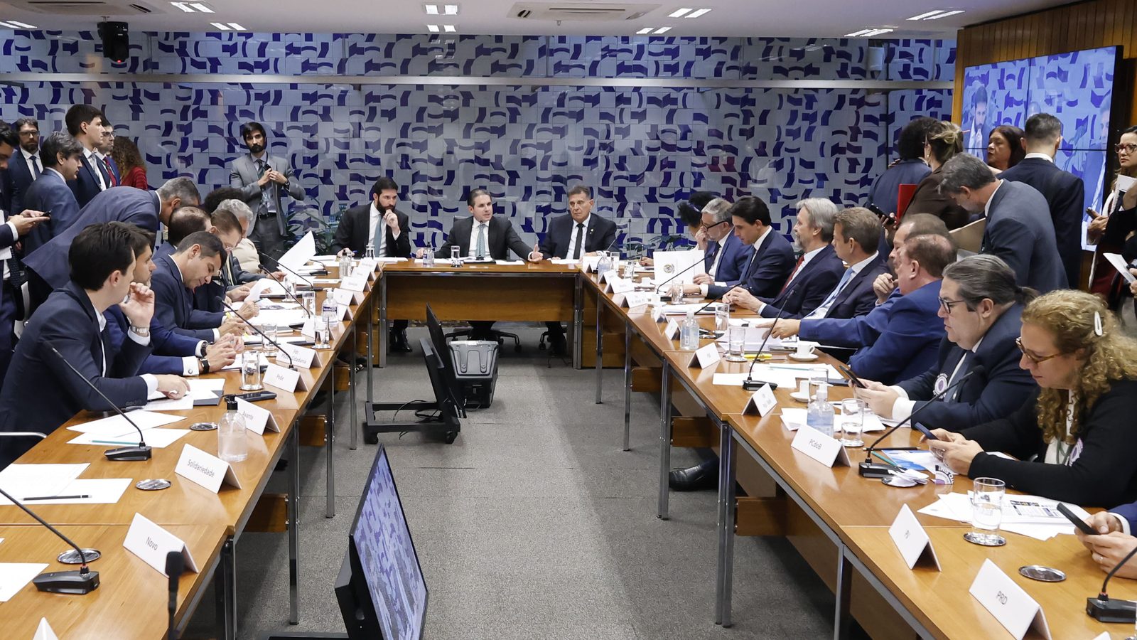 Fotografia. Parlamentares sentados em torno de uma mesa comprida em sala de reuniões da Câmara dos Deputados, alguns conversando entre si, com microfones à frente de cada lugar.