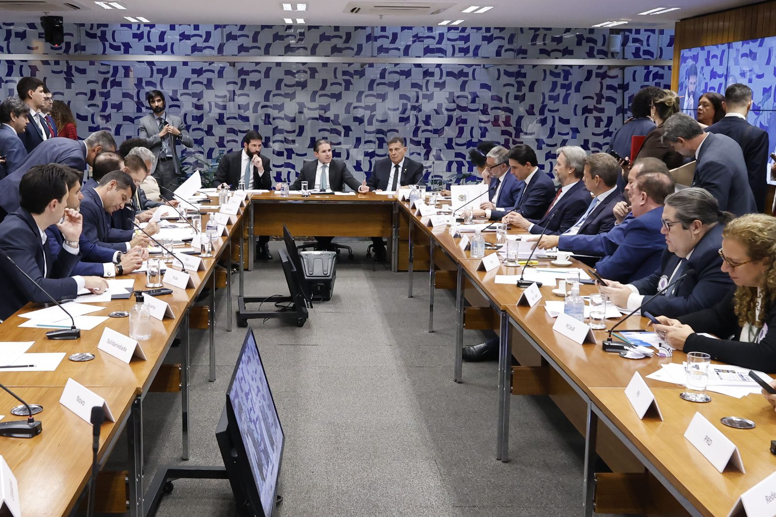 Fotografia. Parlamentares sentados em torno de uma mesa comprida em sala de reuniões da Câmara dos Deputados, alguns conversando entre si, com microfones à frente de cada lugar.