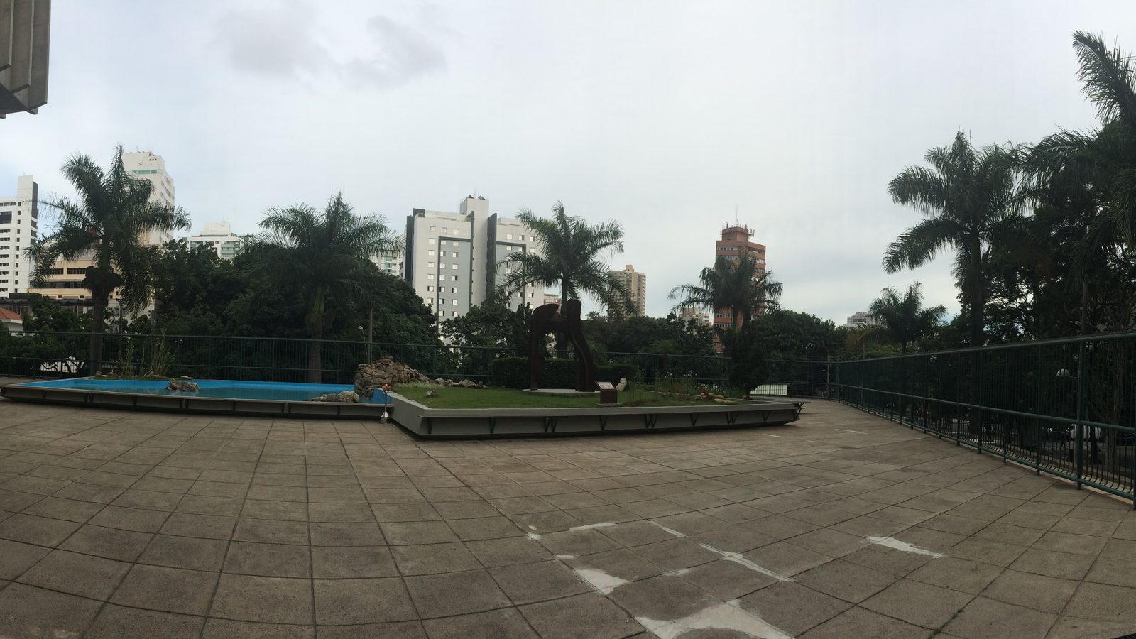 Fotografia panorâmica. Edifício corporativo horizontal de vários andares, com fachada de concreto e faixas de janelas, ocupando um quarteirão em avenida arborizada de Belo Horizonte.