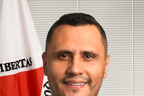 Fotografia. Cleitinho aparece de terno cinza e gravata escura diante de fundo claro e bandeira de Minas Gerais.