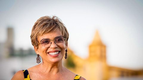 Fotografia. Marília Campos sorri usando óculos, brincos e vestido estampado, com uma paisagem urbana desfocada ao fundo.