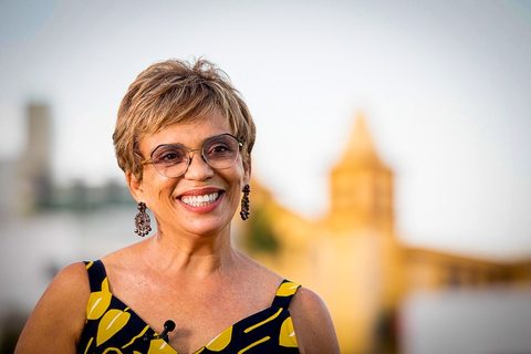 Fotografia. Marília Campos sorri usando óculos, brincos e vestido estampado, com uma paisagem urbana desfocada ao fundo.
